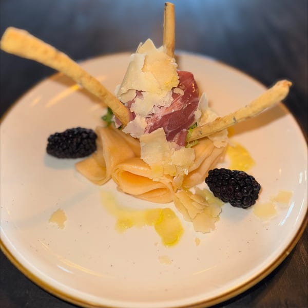 Italian appetizer with prosciutto, shaved parmesan, breadsticks, crepes, and blackberries on a white plate.